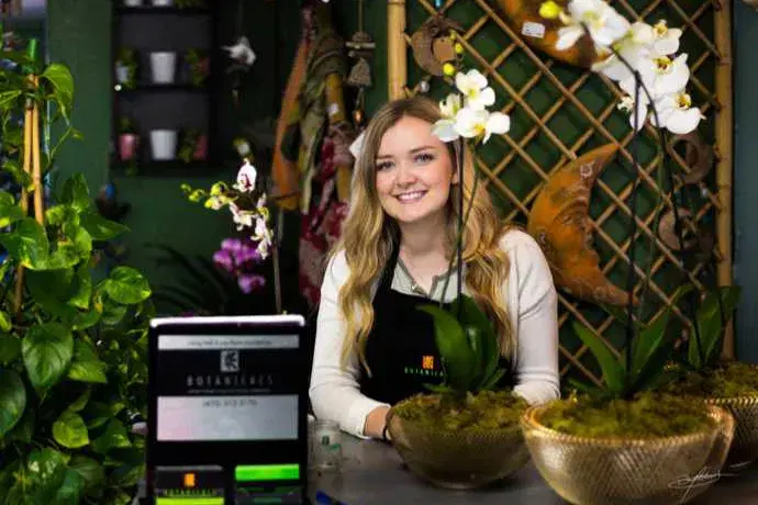 a woman smiling and standing behind retail counter with flowers and plants placed on top