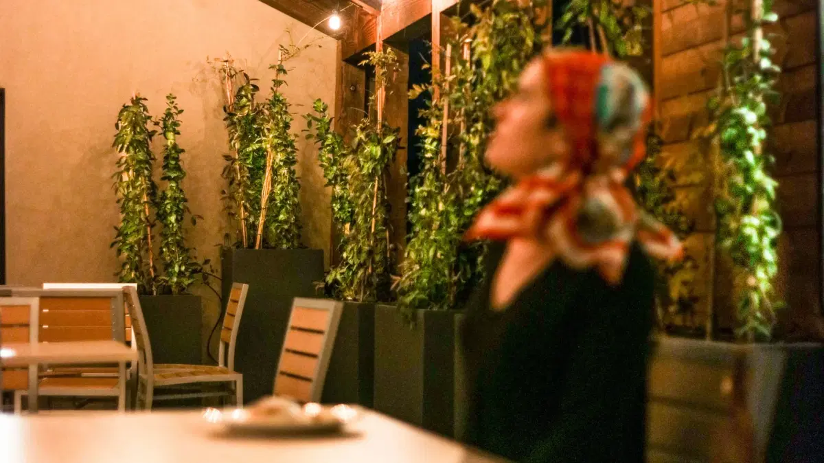 a woman sitting at a table in a restaurant in front of plantscaped pottery